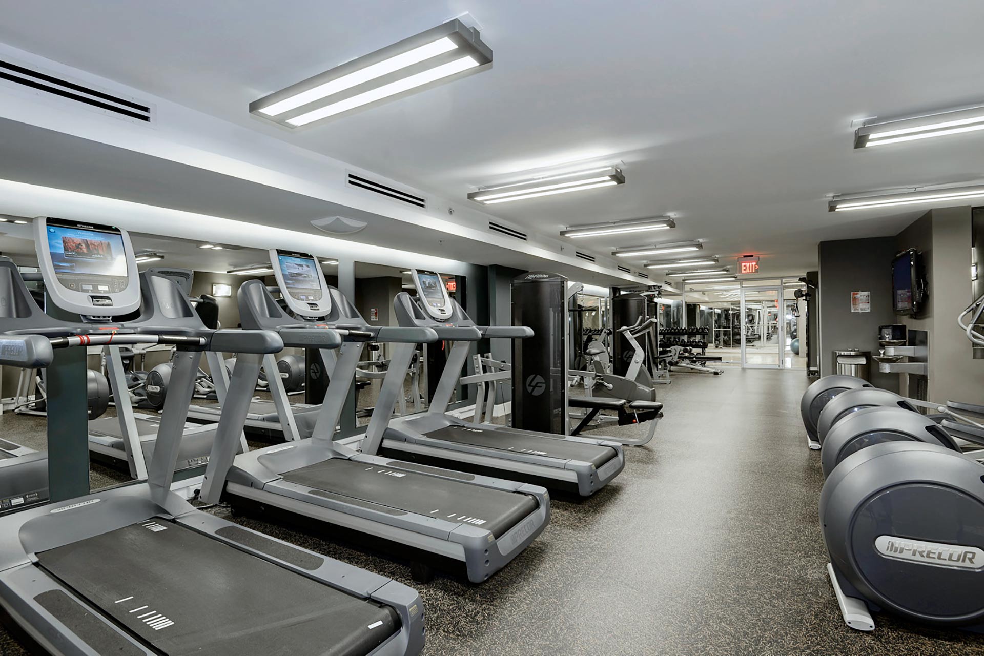 Large fitness room with impact absorbent flooring and cardio equipment along the left, mirrored wall
