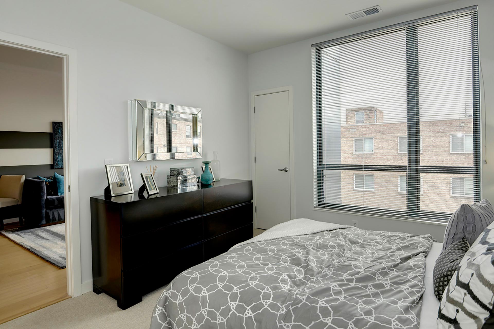 Model unit bedroom with queen bed, dresser, and large window, with door open to the living room on the far left